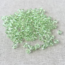 Load image into Gallery viewer, A scattered pile of 6mm Austrian Bicone Crystals - Chrysolite Green with light green faceted surfaces resting on a neutral fabric background.