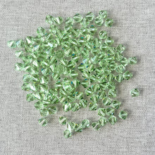 Load image into Gallery viewer, 6mm Austrian Bicone Crystals - Chrysolite Green scattered on a textured gray fabric, showcasing their faceted, translucent light green surfaces ideal for jewelry making.