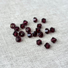 Load image into Gallery viewer, 5mm Burgundy Austrian Bicone Crystals - Burgundy - Package of 20 scattered on a textured neutral fabric background, showcasing their rich deep red color and faceted sparkle.
