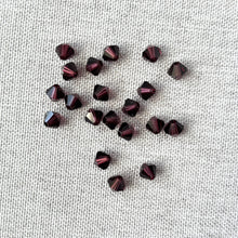 Load image into Gallery viewer, Scattered 5mm Burgundy Austrian Bicone Crystals - Burgundy - Package of 20 on a textured light fabric background, showcasing their deep red color and faceted design.