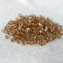 Load image into Gallery viewer, A close-up view of numerous faceted 6mm Light Colorado Topaz Austrian Bicone Crystals - Light Colorado Topaz Brown scattered on a neutral fabric surface.