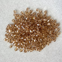 Load image into Gallery viewer, 6mm Light Colorado Topaz Austrian Bicone Crystals - Light Colorado Topaz Brown scattered on a light fabric background, showcasing their faceted shine and warm brown color.