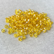 Load image into Gallery viewer, A close-up of multiple 6mm Light Topaz Austrian Bicone Crystals - Light Topaz Yellow arranged on a light fabric surface, showcasing their faceted, sparkling texture.