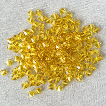 Load image into Gallery viewer, A close-up view of numerous 6mm Light Topaz Austrian Bicone Crystals - Light Topaz Yellow scattered on a light fabric background, showcasing their bright yellow faceted surfaces.
