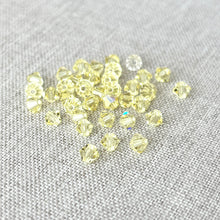 Load image into Gallery viewer, 4mm Jonquil CRYSTALLIZED™ Bicone Crystals - Jonquil Yellow - Pack of 37 scattered on a textured light gray fabric background, showing their faceted, translucent yellow design.