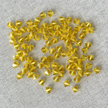 Load image into Gallery viewer, Scattered 5mm Light Topaz Austrian Bicone Crystals in Light Topaz Yellow on a textured surface, showcasing the vibrant color and faceted design of the pack of 100.