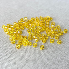 Load image into Gallery viewer, 5mm Light Topaz Austrian Bicone Crystals - Light Topaz Yellow - Pack of 100 scattered on a neutral fabric background, showcasing their faceted, sparkling surfaces ideal for jewelry making.