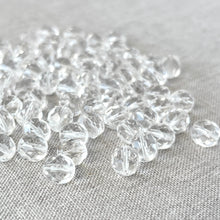 Load image into Gallery viewer, A close-up of multiple 8mm Crystal Clear Austrian Round Crystals - Crystal Clear scattered on a textured light fabric surface, showcasing their faceted, transparent design.