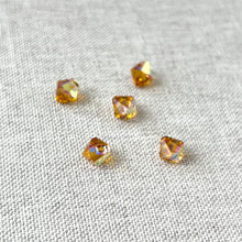 Load image into Gallery viewer, 6mm Topaz AB Austrian Bicone Drop Crystals - Topaz AB Brown - Top Drilled scattered on a textured off-white fabric background, showing their faceted, reflective surfaces and warm brown hues.