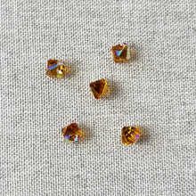 Load image into Gallery viewer, Close-up of five 6mm Topaz AB Austrian Bicone Drop Crystals - Topaz AB Brown - Top Drilled scattered on a textured beige fabric background, showing their facet brilliance and iridescent hues.