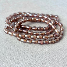 Load image into Gallery viewer, Close-up of two 16 inch strands of Rootbeer Glass Oval Beads - Pack of 2 - 16 inch strands - Translucent Glass with a warm brown translucent finish on a neutral fabric background.