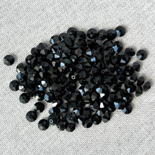 Load image into Gallery viewer, A pile of jet black faceted rondelle crystals sparkling with reflective surfaces on a light fabric background, showcasing the Jet Black CRYSTALLIZED™ Austrian Faceted Rondelle Crystal - 6x4mm - Cut 5305.