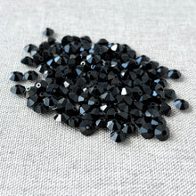Load image into Gallery viewer, A close-up view of multiple Jet Black CRYSTALLIZED™ Austrian Faceted Rondelle Crystal - 6x4mm - Cut 5305 beads with a shiny, faceted surface resting on a neutral fabric background.