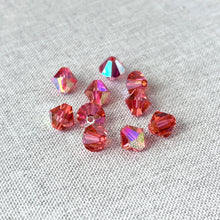 Load image into Gallery viewer, Close-up of scattered 8mm Padparadscha AB - Austrian Bicone Crystals in vibrant pink-orange hues with iridescent reflections on a textured gray fabric background.