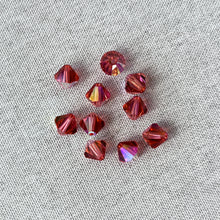 Load image into Gallery viewer, A collection of 8mm Padparadscha AB - Austrian Bicone Crystals with a vibrant pinkish-orange hue and faceted surfaces, arranged on a neutral fabric background.