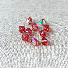 Load image into Gallery viewer, A small cluster of vibrant pinkish-orange 8mm Padparadscha AB - Austrian Bicone Crystals with shimmering faceted surfaces on a light textured fabric background.