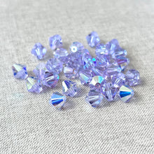 Load image into Gallery viewer, A scattered pile of 8mm Austrian Bicone Crystals - AB Alexandrite - Finish AB with a light purple hue and reflective sparkling facets on a beige fabric background.