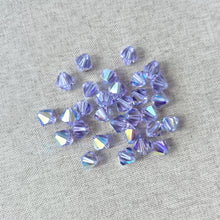 Load image into Gallery viewer, 8mm Austrian Bicone Crystals - AB Alexandrite - Finish AB scattered on a textured light gray fabric, showcasing their faceted purple and iridescent colors for jewelry making.