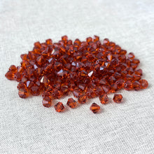 Load image into Gallery viewer, A pile of 6mm Austrian Bicone Crystals - Indian Red scattered on a light fabric surface, showing their faceted, translucent deep red color and sparkle.