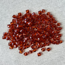 Load image into Gallery viewer, A close-up of sparkling 6mm Austrian Bicone Crystals - Indian Red scattered on a textured light fabric background, showcasing their rich red color and faceted cuts.