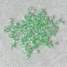 Load image into Gallery viewer, 4mm CRYSTALLIZED™ Austrian Bicone Crystals - Chrysolite Green - Pkg of 136 scattered on a textured light gray fabric background, showcasing their faceted, sparkling green appearance.