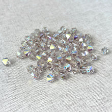 Load image into Gallery viewer, A scattered pile of sparkling translucent crystals with an iridescent finish on a light textured fabric surface, showcasing 6mm Cantaloupe AB Austrian Bicone Crystals - AB Cantaloupe.