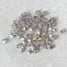 Load image into Gallery viewer, Scattered 6mm Cantaloupe AB Austrian Bicone Crystals - AB Cantaloupe with iridescent reflections on a light textured fabric background.