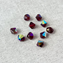 Load image into Gallery viewer, A collection of 8mm Garnet AB Austrian Bicone Crystals - Garnet AB Red scattered on a light textured fabric surface, showing deep red facets with iridescent blue and green reflections.
