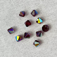 Load image into Gallery viewer, Scattered 8mm Garnet AB Austrian Bicone Crystals - Garnet AB Red with deep red color and reflective iridescent coating on a light textured fabric background.