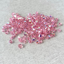 Load image into Gallery viewer, 5mm and 6mm Light Rose AB - Austrian Bicone Crystals - Light Rose - AB Finish - Package of 150 scattered on a light fabric background, showing their faceted, shimmering surfaces.