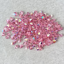 Load image into Gallery viewer, 5mm and 6mm Light Rose AB - Austrian Bicone Crystals - Light Rose - AB Finish - Package of 150 shiny faceted crystals with iridescent rainbow reflections on a neutral fabric background
