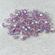 Load image into Gallery viewer, A scattered pile of 6mm Lt Amethyst AB Austrian Bicone Crystals - Light Amethyst - AB Finish beads on a neutral fabric surface, showing their faceted sparkle and iridescent coating.