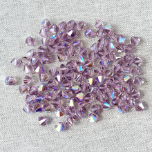 Load image into Gallery viewer, Scattered 6mm Lt Amethyst AB Austrian Bicone Crystals - Light Amethyst - AB Finish with reflective, faceted surfaces on a light fabric background.