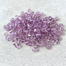 Load image into Gallery viewer, A pile of 6mm Lt Amethyst Austrian Bicone Crystals - Light Amethyst in a soft purple shade displayed on a textured fabric surface.