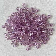 Load image into Gallery viewer, 6mm Lt Amethyst Austrian Bicone Crystals - Light Amethyst beads scattered on a light fabric background, showcasing their faceted cuts and soft purple color.