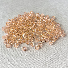 Load image into Gallery viewer, Close-up of numerous 6mm Light Peach - Austrian Bicone Crystals - Light Peach scattered on a light fabric surface, showcasing their faceted cut and soft peach color.