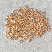 Load image into Gallery viewer, A close-up view of multiple 6mm Light Peach - Austrian Bicone Crystals - Light Peach scattered on a neutral fabric background, showcasing their faceted, sparkling surfaces.