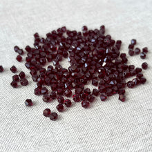 Load image into Gallery viewer, 4mm Garnet CRYSTALLIZED™ Austrian Bicone Crystals - Garnet Red scattered on a light fabric background showing their deep red color and faceted design.