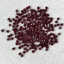 Load image into Gallery viewer, 4mm Garnet CRYSTALLIZED™ Austrian Bicone Crystals - Garnet Red scattered on a light fabric surface, showing their deep red color and faceted sparkle.