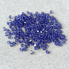 Load image into Gallery viewer, 4mm Tanzanite - Austrian Bicone Crystals - Tanzanite Purple - Package of 200 scattered on a light fabric surface showcasing their faceted, shiny texture for jewelry making.