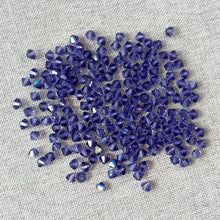 Load image into Gallery viewer, 4mm Tanzanite - Austrian Bicone Crystals - Tanzanite Purple - Package of 200 scattered on a textured light gray fabric background, showcasing their deep purple faceted surfaces and vibrant sparkle.