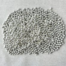 Load image into Gallery viewer, A close-up view of numerous silver-plated corrugated round spacer beads scattered on a light fabric surface, showcasing the 6mm size and texture from the Spacer Beads - Metal Beads - Corrugated Rounds - Silver Plated - 6mm - 8 oz bag package.