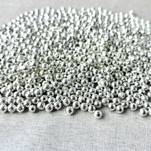 Load image into Gallery viewer, Close-up view of numerous small, silver-plated corrugated round spacer beads scattered on fabric, matching Spacer Beads - Metal Beads - Corrugated Rounds - Silver Plated - 6mm - 8 oz bag package.