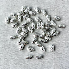 Load image into Gallery viewer, Swirl Diamond Cut Silver Plated Cylinder Beads Oval - Spacer Beads Metal Beads - Package of 48 scattered on a textured fabric surface, showing their shiny silver finish and detailed swirl cut design.