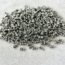 Load image into Gallery viewer, Pile of small Antique Silver - 7mm x 3mm - Tube with Beaded Flower beads on a textured fabric background, ideal for jewelry making and craft projects.